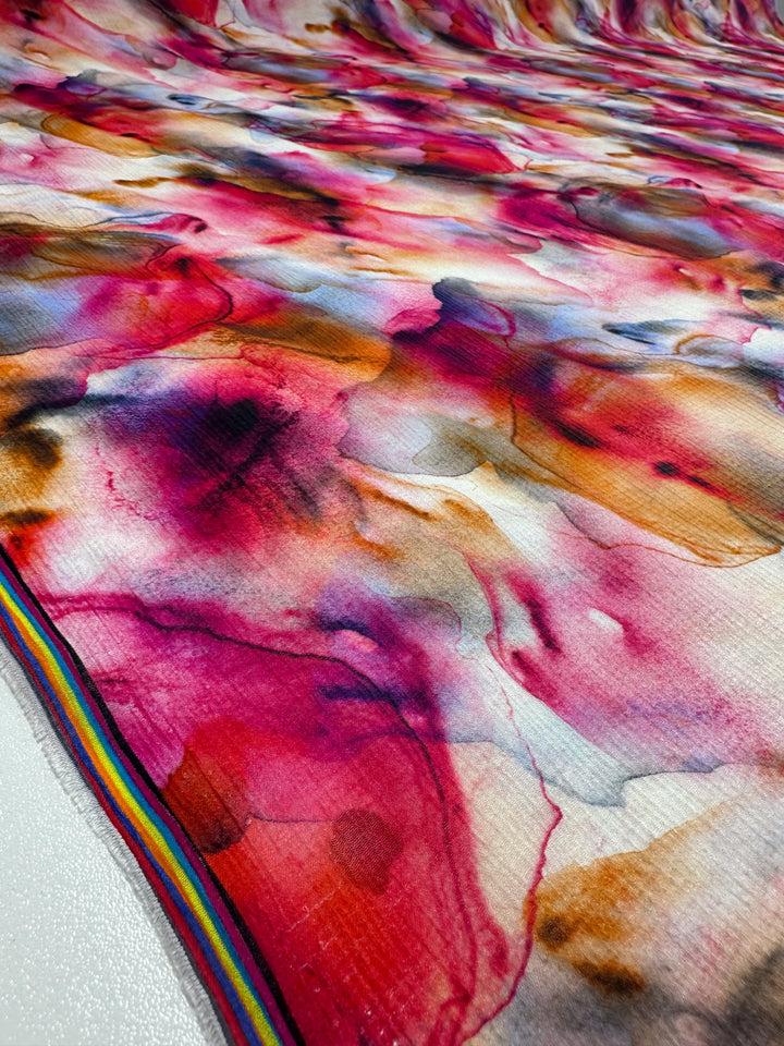 Super Cheap Fabrics' "Designer Cotton - Zabaleta - 150cm" features a vibrant abstract pattern in swirling pink, red, and orange hues. It has a small rainbow stripe along the edge for contrast. The luxury fabric is perfect for sewing projects with its smooth, slightly glossy texture.