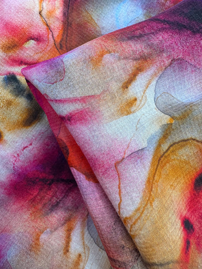 The Designer Cotton - Zabaleta by Super Cheap Fabrics features a close-up of breathable fabric with a vibrant watercolor pattern in swirling blends of pink, red, orange, and yellow hues. This abstract artistic design has a soft texture, ideal for luxury sewing projects. The fabric measures 150cm wide.