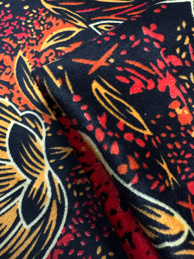 Close-up of Super Cheap Fabrics' DD Velvet - Ember Bloom (150cm), showcasing its vibrant red, yellow, orange, and black abstract design with stylized leaves; the folded polyester fabric reveals rich texture and intricate details.