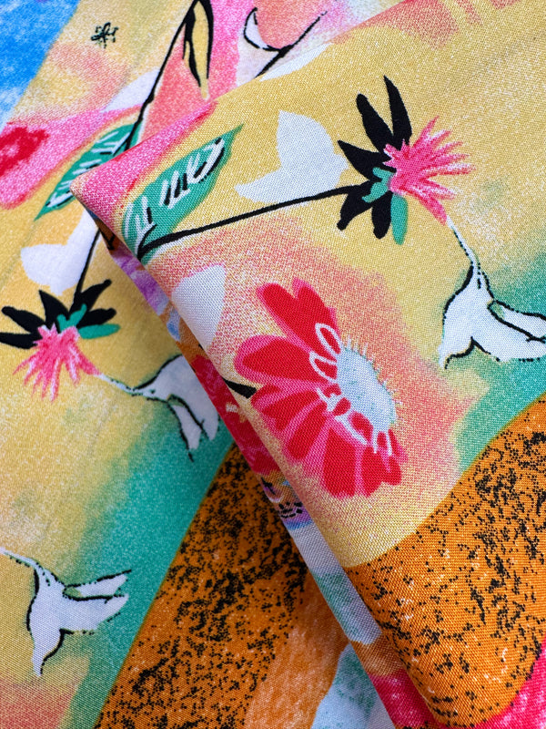 A close-up of Super Cheap Fabrics’ Designer Rayon - Wildframe (145cm) shows vibrant abstract floral and bird patterns in pink, white, black, green, yellow, orange, and turquoise with a soft texture and striking colors.