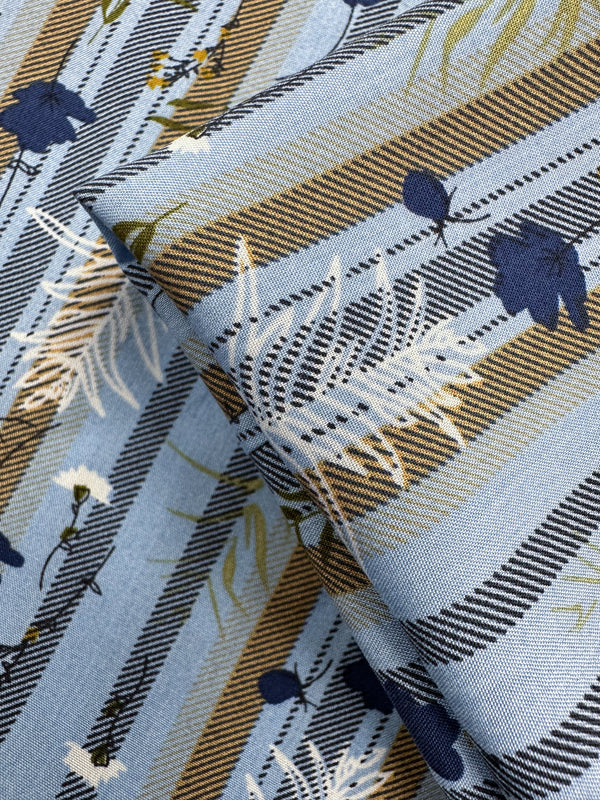 Designer Rayon - Fieldstripe Bloom by Super Cheap Fabrics features blue rayon with diagonal brown, white, and black stripes, plus a white and navy flower and leaf pattern. Slightly folded to show texture. Width: 145cm.