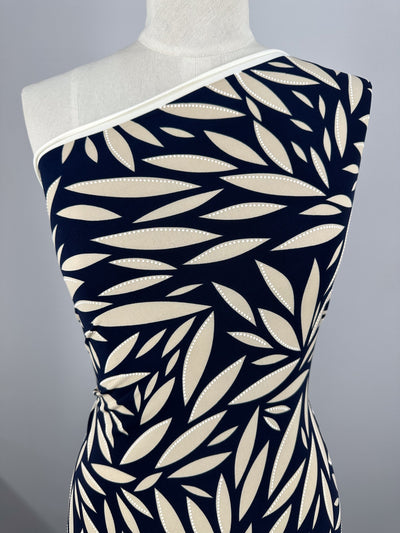 A mannequin wears a fitted one-shoulder dress made from Super Cheap Fabrics' Textured Lycra - Prism Leaf (150cm), showcasing a bold navy and beige leaf design with white dotted outlines and white trim at the neckline.