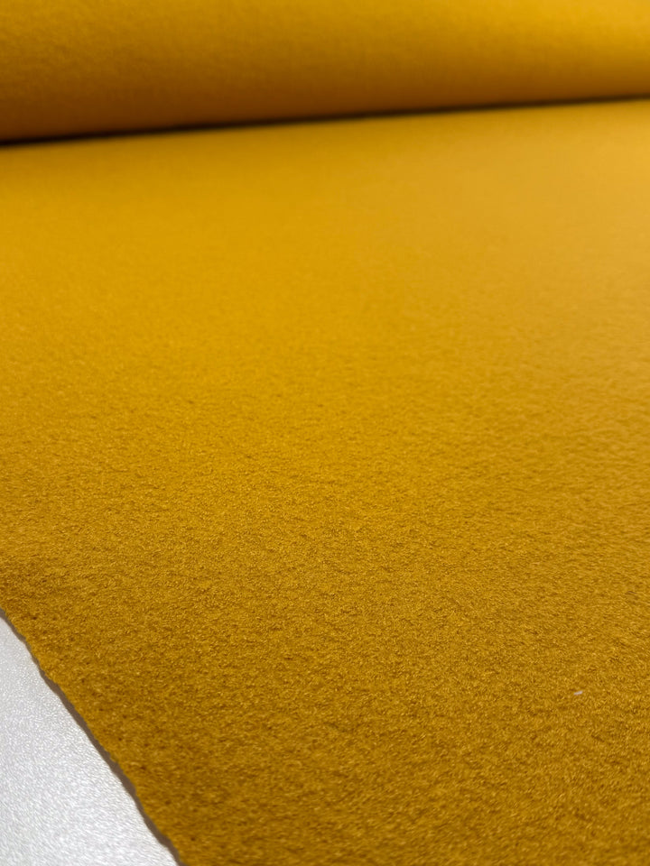 A close-up of Super Cheap Fabrics' Boiled Wool - Mango Mojito (150cm), showing its soft, felt-like texture and vibrant mustard yellow color, partially unrolled on a flat surface.