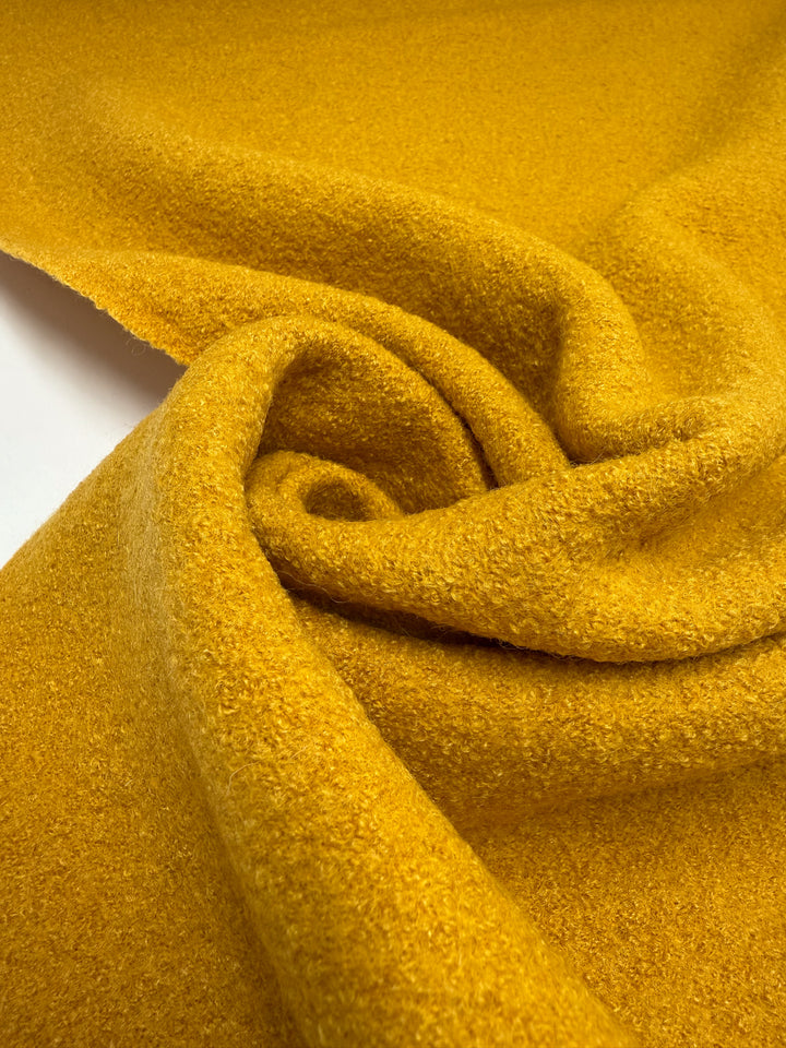 A close-up of Super Cheap Fabrics' Boiled Wool - Mango Mojito (150cm) displays its thick, textured surface in a loose spiral, highlighting the fabric's soft folds and vibrant mustard yellow hue.