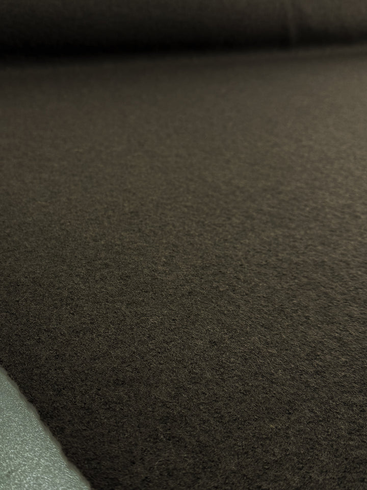 A close-up of Super Cheap Fabrics' Boiled Wool - Forest Night (152cm), dark brown and textured with a slightly fuzzy finish, laid flat. The bottom left shows a metallic or plastic edge—ideal for extra heavy weight winter garments.