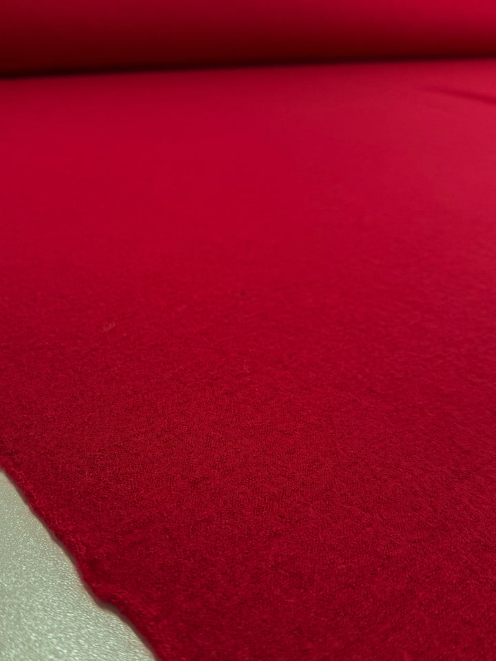 A close-up of Super Cheap Fabrics' Boiled Wool - Crimson - 145cm rolled out, highlights its vibrant red color, soft wool texture, and smooth, slightly fuzzy finish with the roll visible at the top.