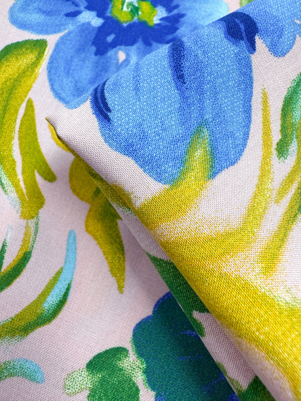 A close-up of Super Cheap Fabrics' Designer Rayon - Bluebell Blush (145cm), showcasing its vibrant blue and green floral print with yellow accents on a light background, and subtle folds that highlight the fabric’s soft texture.