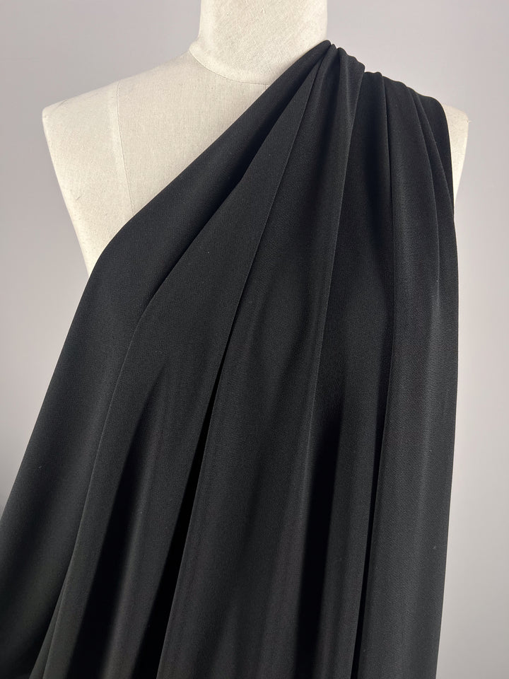 The Crepe Lycra in black by Super Cheap Fabrics is elegantly draped over one shoulder of a mannequin, creating a flowing and sophisticated look against a neutral backdrop.