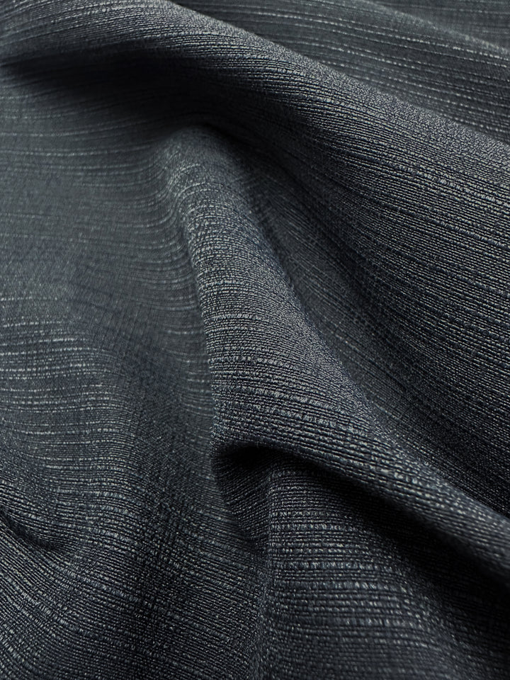 Close-up of Super Cheap Fabrics' Textured Bengaline in Textured Indigo, 145cm wide. The medium-weight, dark gray stretch fabric features a ribbed surface and is gently draped to create soft folds and a subtle sheen highlighting the weave pattern.