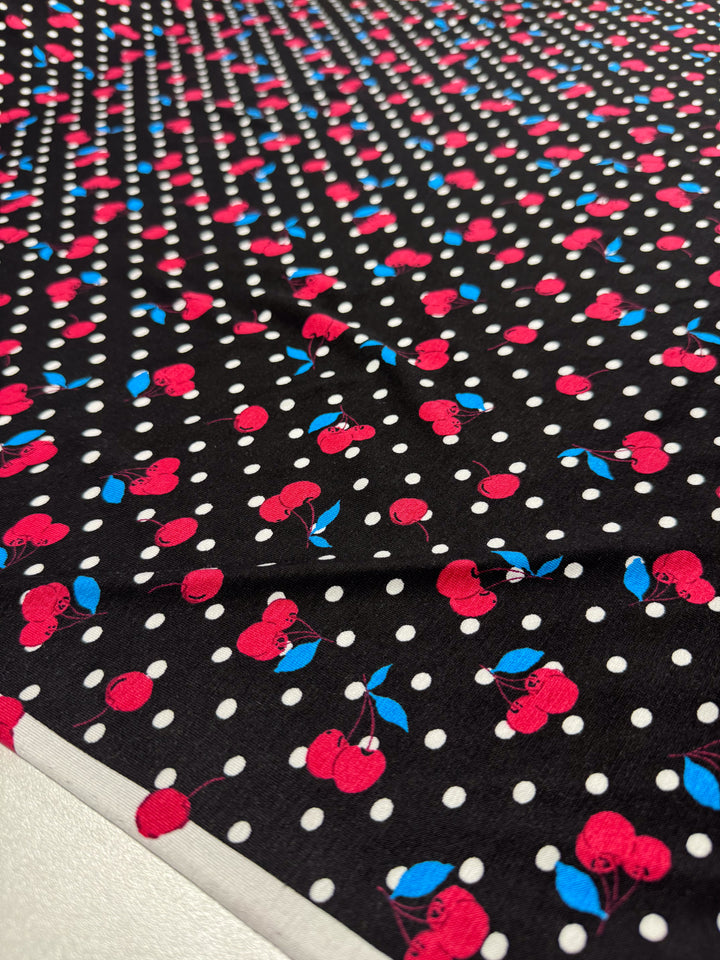 The Super Cheap Fabrics "Printed Cotton Lycra - Cherry Black - 150cm" features medium-weight lycra fabric with bright red cherries, blue leaves, and white polka dots on a black background. Ideal for versatile clothing, this eye-catching material is displayed across a surface.