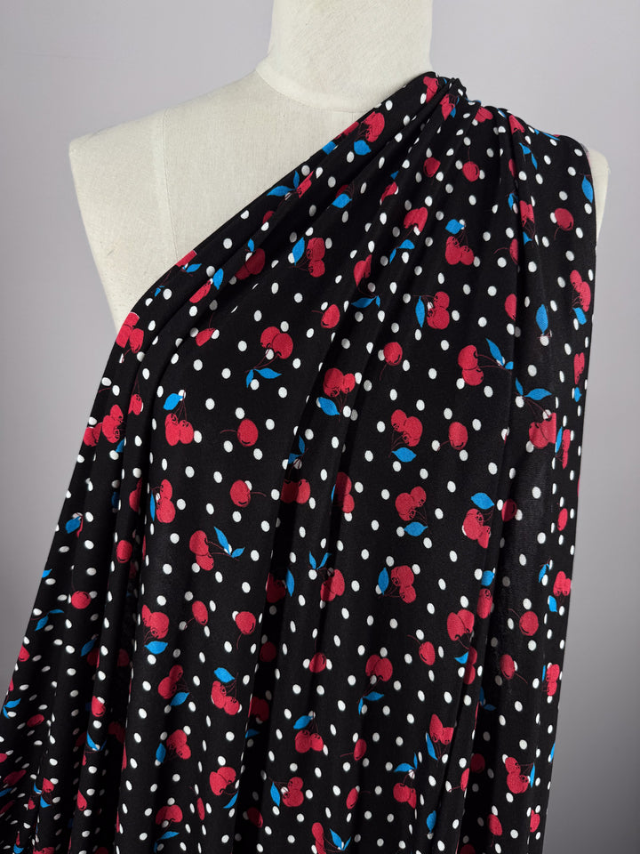 A mannequin displayed on a gray background wears Printed Cotton Lycra - Cherry Black from Super Cheap Fabrics. The medium-weight fabric features a black base with white polka dots, red cherries, and blue leaves, ideal for versatile clothing designs.