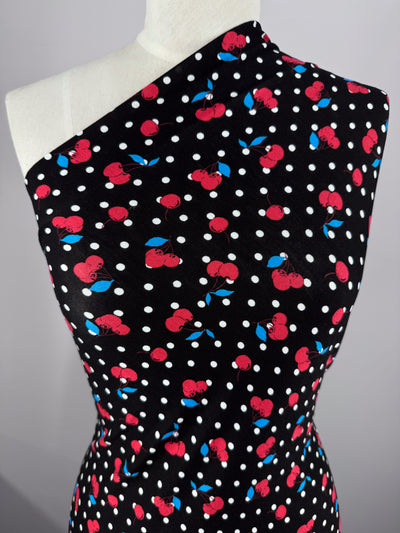 The Cherry Black printed cotton lycra from Super Cheap Fabrics drapes elegantly on a mannequin, featuring a one-shoulder design. This fabric showcases black with white polka dots and red cherries, in medium weight.
