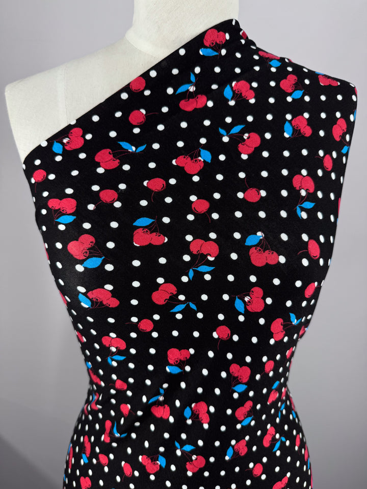The Cherry Black printed cotton lycra from Super Cheap Fabrics drapes elegantly on a mannequin, featuring a one-shoulder design. This fabric showcases black with white polka dots and red cherries, in medium weight.