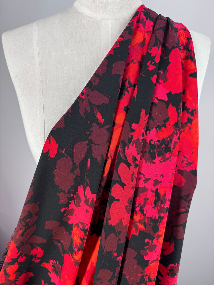 A close-up of a mannequin showcasing Super Cheap Fabrics' "Printed Crepe - Burn - 150cm," featuring a vibrant red and black floral pattern. The asymmetrical design across the torso offers a striking contrast against the plain background, ideal for fashion garments.