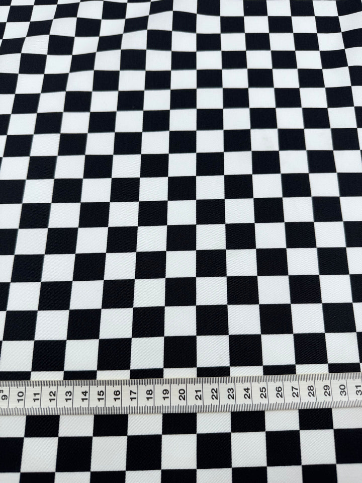 The Cotton Drill - Checkmate - 148cm by Super Cheap Fabrics is a lightweight black and white fabric with evenly spaced checkerboard squares. Featuring a durable diagonal weave, its measurements are shown in centimeters with a metal ruler at the bottom for scale.