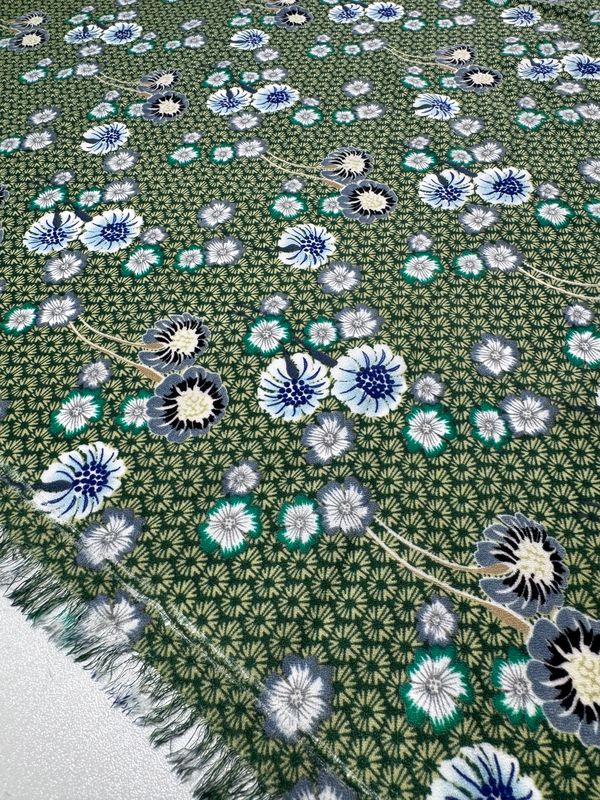 The Printed Crepe - Botan by Super Cheap Fabrics is a 148cm wide fabric featuring a green base with a floral pattern in white, blue, and black. It includes slight fraying on the edges and small geometric shapes in the background for added texture and elegance.