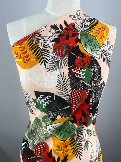 The Super Cheap Fabrics' Printed Crepe - Jungle Sunset - 140cm drapes over the mannequin, showcasing a luxurious pattern of red, yellow, green, and black abstract botanical leaves against a beige background. The lightweight fabric is wrapped elegantly, highlighting its colorful design.
