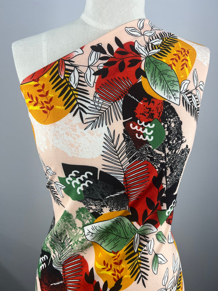 The Super Cheap Fabrics' Printed Crepe - Jungle Sunset - 140cm drapes over the mannequin, showcasing a luxurious pattern of red, yellow, green, and black abstract botanical leaves against a beige background. The lightweight fabric is wrapped elegantly, highlighting its colorful design.